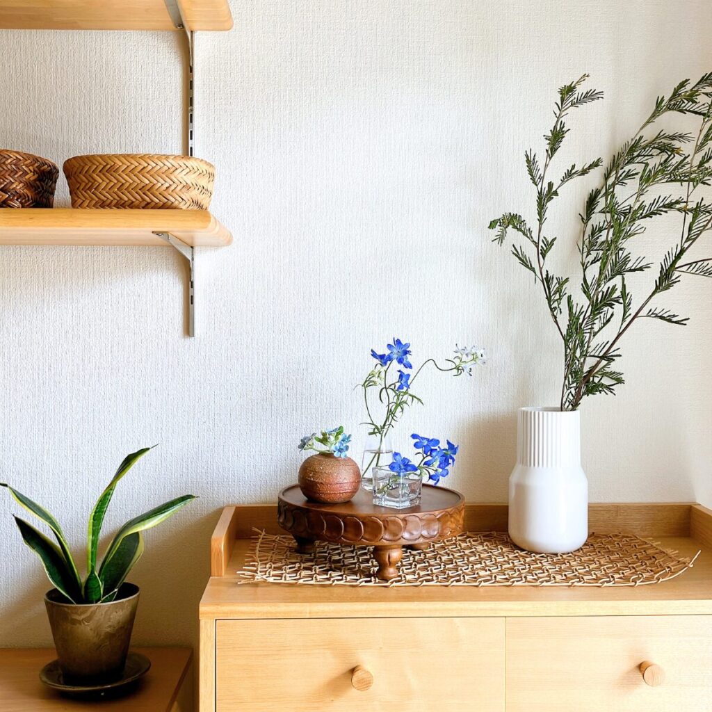 A wide interior shot of a room corner featuring a blue flower arrangement on a compote, framed by Acacia branches and houseplants