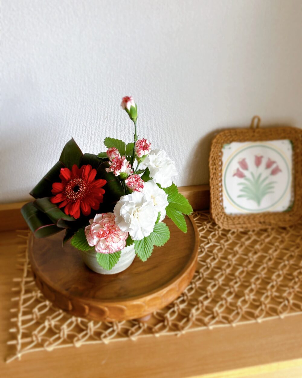 A red gerbera framed by dracaena leaves and white carnations in a plump Kasama pottery jar on a wooden compote.