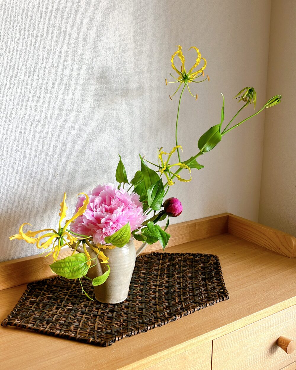 A pink peony and yellow gloriosa spreading left and right in a Kasama pottery pitcher on a wooden shelf.