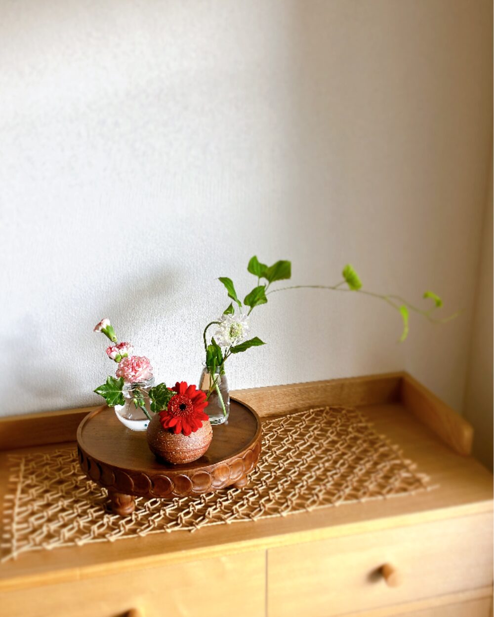 A red gerbera, white carnations, and scabiosa arranged across three small ceramic and glass vases on a wood compote.