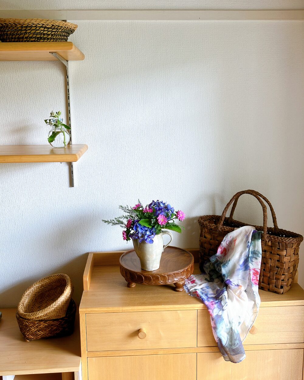 A wide view of the ceramic pitcher with flowers, with a basket bag and a scarf placed right next to it.