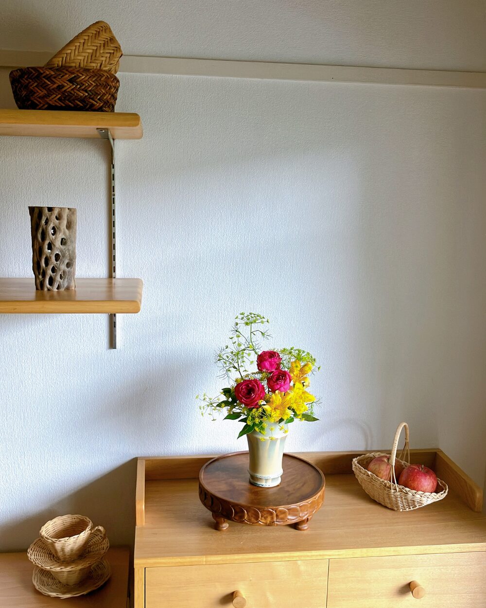 Deep pink rose in a ceramic vessel with a basket of apples on a shelf