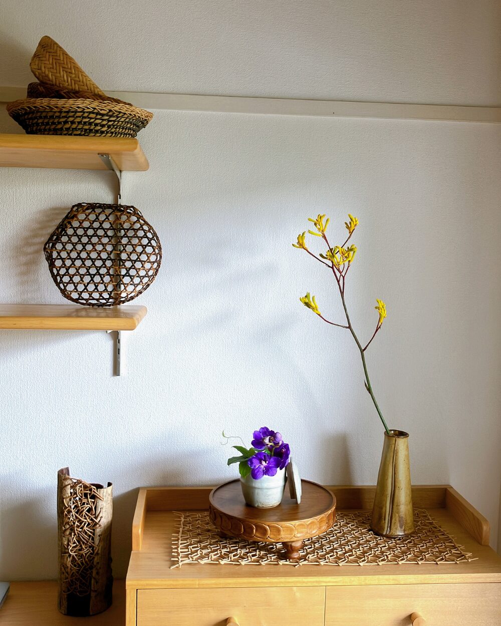 A Vanda orchid in a ceramic jar on a wood compote, styled with a woven basket and a rattan lampshade.
