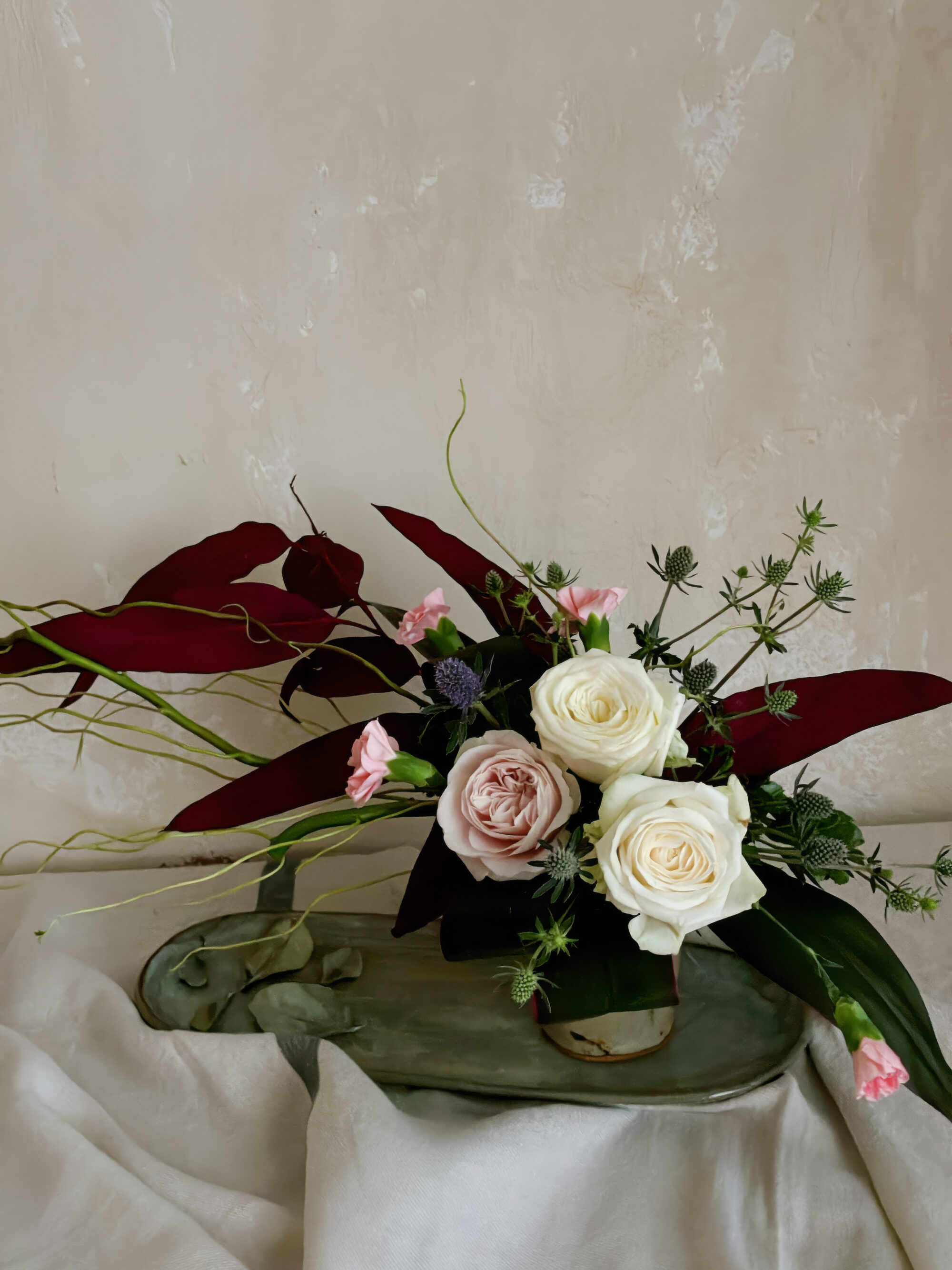 An arrangement of roses styled in a Japanese pottery beer cup, featuring a strong contrast between the bright blossoms and deep-toned preserved eucalyptus leaves.