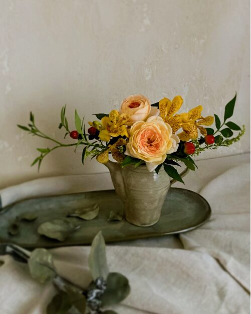 An art poster style photograph of pale orange roses and yellow mokara orchids in a Japanese pottery pitcher, featuring a minimalist aesthetic with strong natural light.