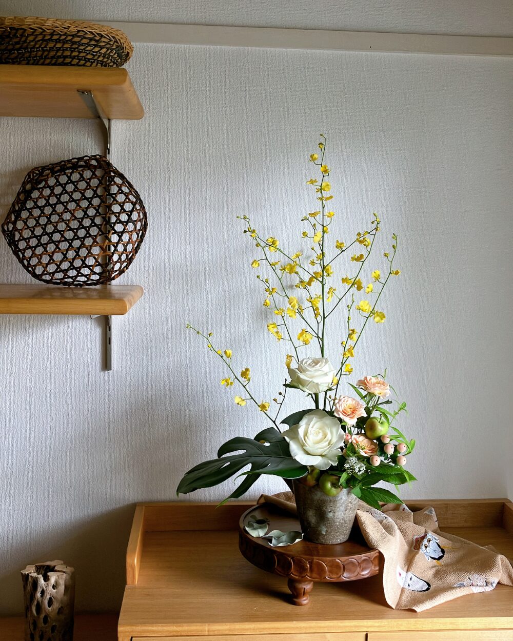 A long shot of the vertical orchid arrangement on a shelf corner styled with a woven basket and a beige furoshiki cloth.