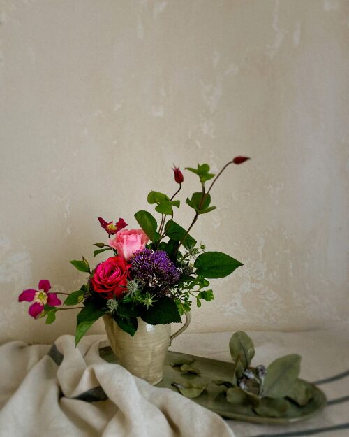 A moody, fine-art photograph of deep pink roses and a gray ceramic pitcher set against a deep shadowed backdrop, highlighting the texture of the glaze and petals.