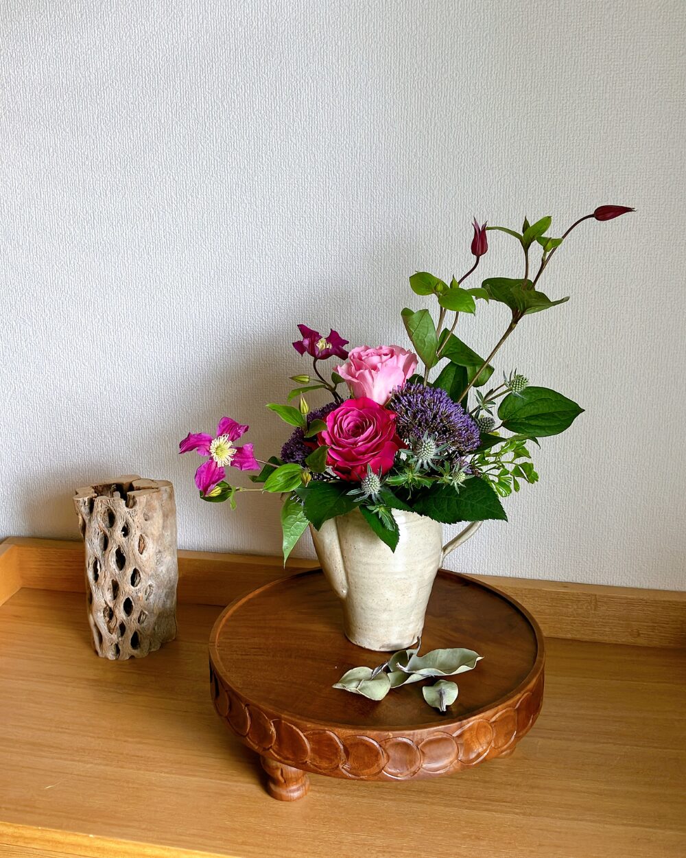 A gray ceramic pitcher containing dark pink and pale pink roses, styled with fluid clematis branches and purple trachelium flowers, viewed from the front.