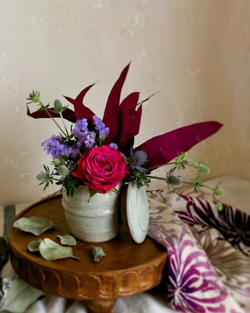 An arrangement focusing on a single deep pink rose within a traditional lidded jar.