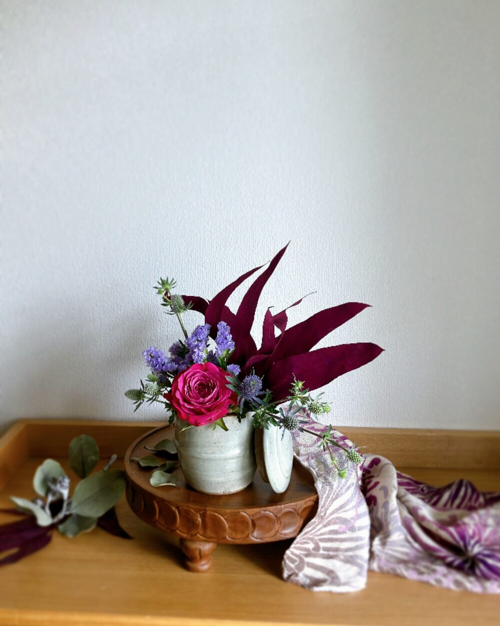 A single deep pink rose in a lidded ceramic jar surrounded by pale purple statice and dark pink preserved eucalyptus, resting on a wooden compote with a striped furoshiki cloth, viewed from the front.
