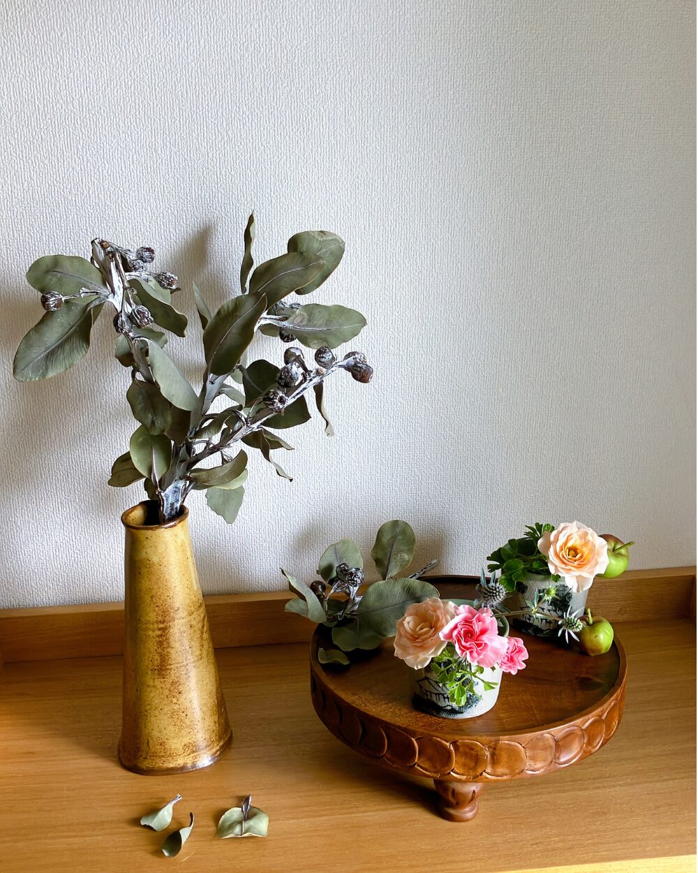 A pink carnation and pale orange spray roses in a Japanese soba choko cup on a wood compote, viewed from the front.