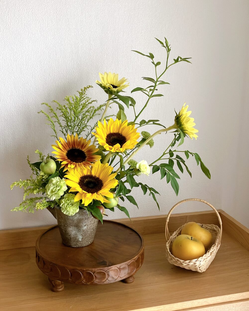 A front view of a flowing sunflower arrangement with yellowish-green carnations and small apples in a dark-colored ceramic vessel.