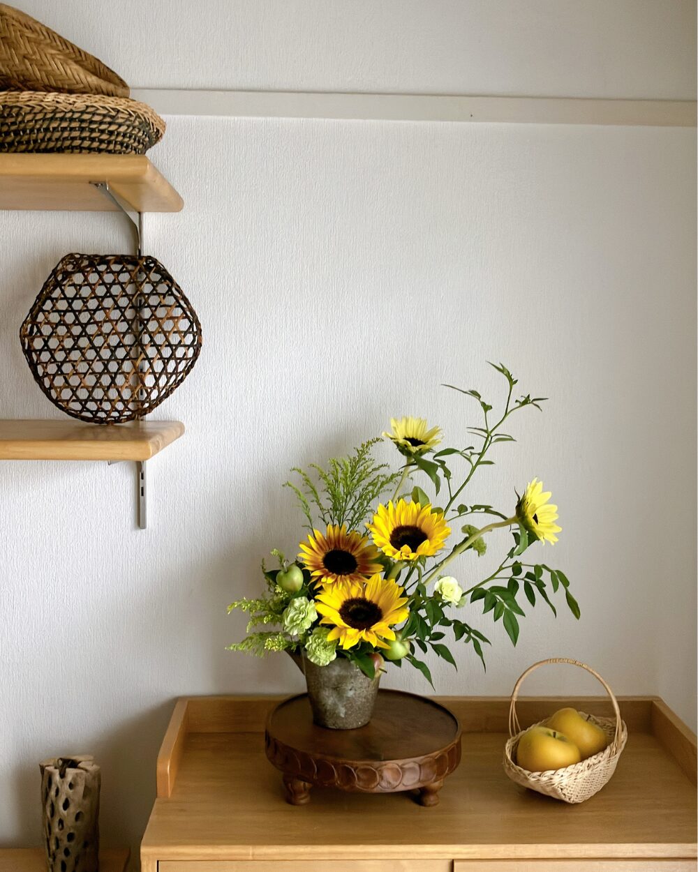 A wide interior view of a calm wooden shelf styled with a dark ceramic flower vessel, a small basket, and yellow apples.
