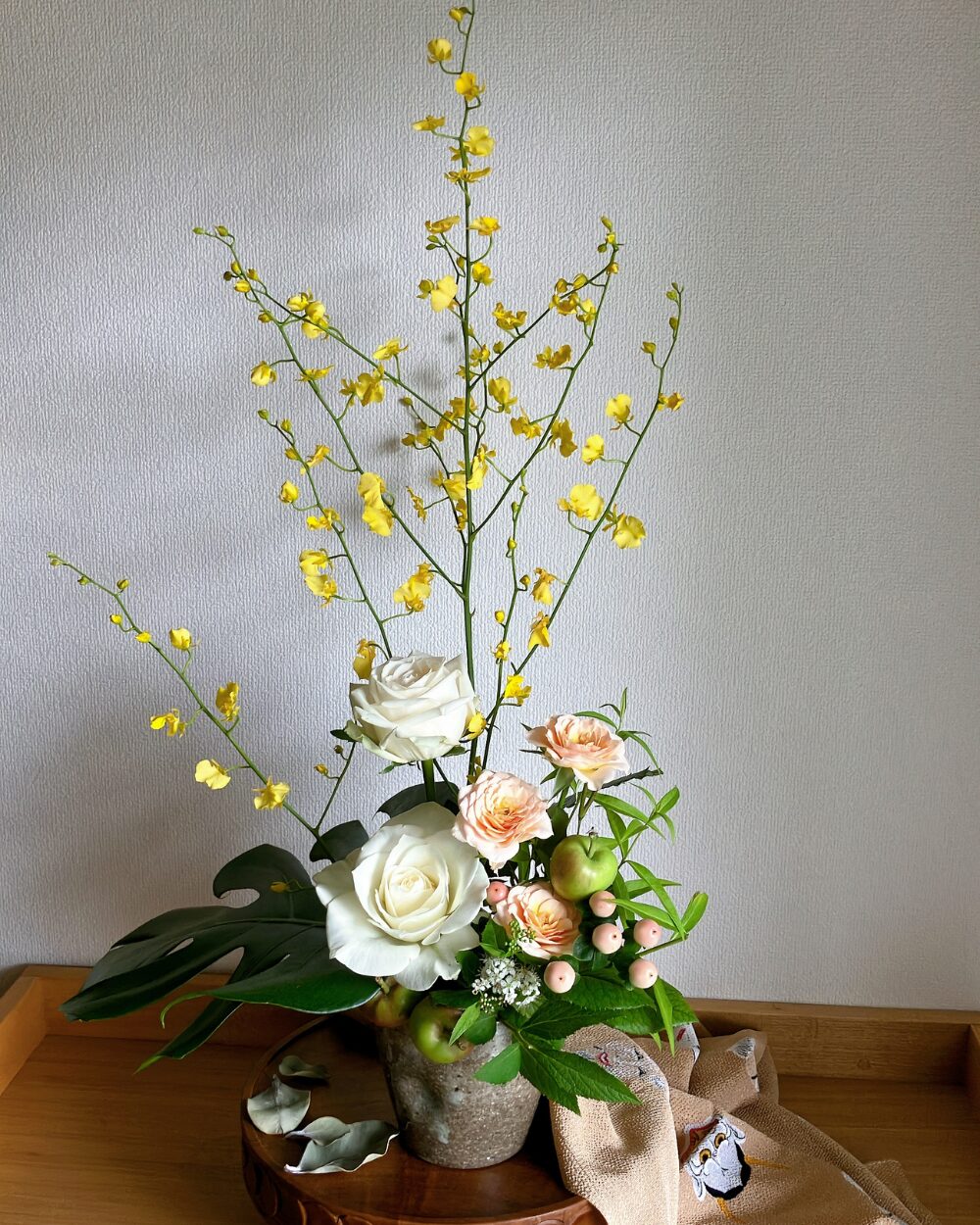 An arrangement of a white rose, yellow oncidium orchids, and pale orange spray roses in a wide Japanese pottery vessel, viewed from the front.