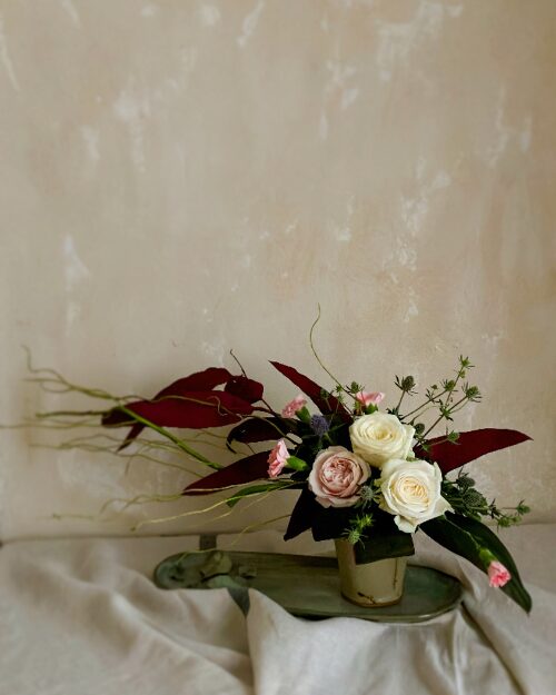 An arrangement of roses styled in a Japanese pottery beer cup, featuring a strong contrast between the bright blossoms and deep-toned preserved eucalyptus leaves.