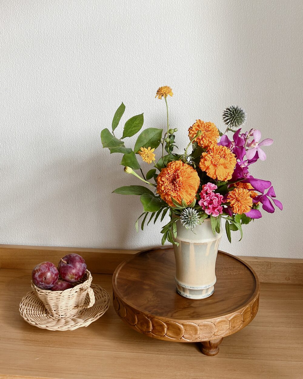 A frontal view of marigolds, mokara, and cockscomb arranged in a tall Japanese ceramic vase with plums placed alongside on a shelf.