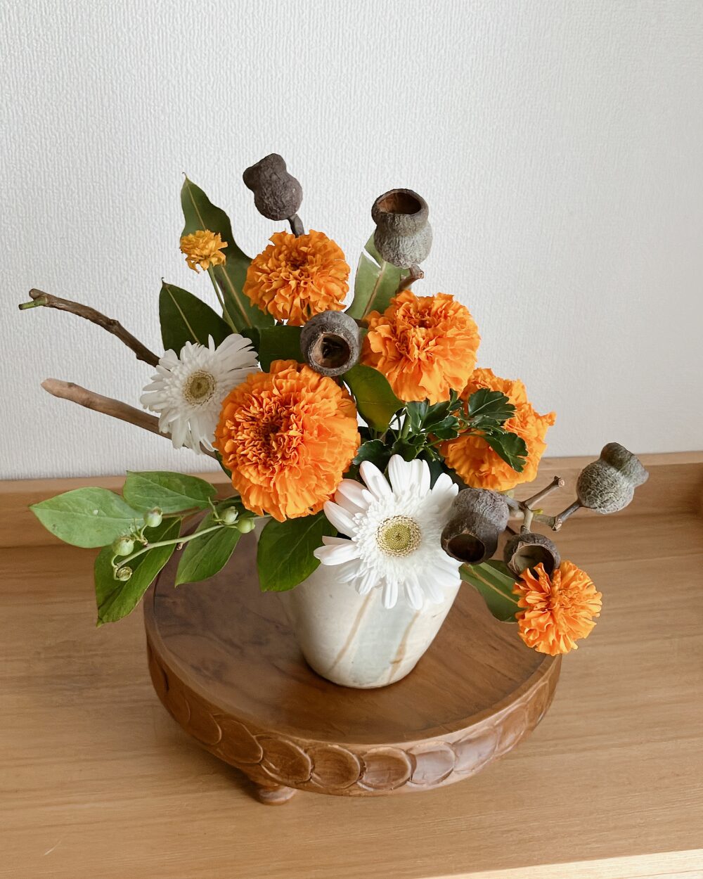 An angled high-angle shot of the arrangement with white gerberas, marigolds, and silver bellgum nuts in a Japanese ceramic vase.