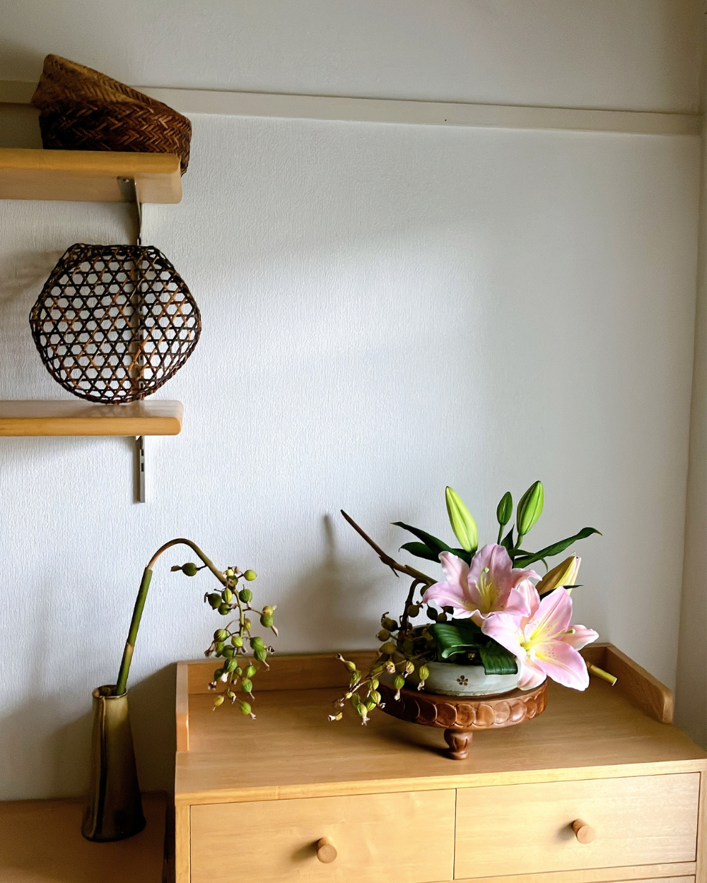 Pink lily blooms and a shell ginger branch arranged on a low Japanese ceramic plate displayed on a wooden shelf.