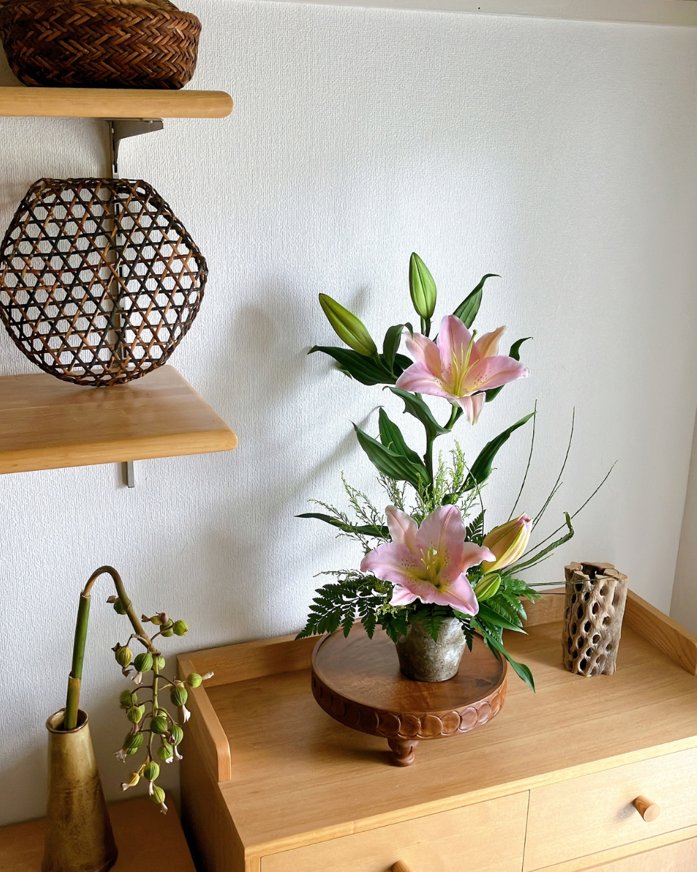 A single tall pink lily arranged in a Japanese ceramic vase on a styled interior shelf with green foliage.